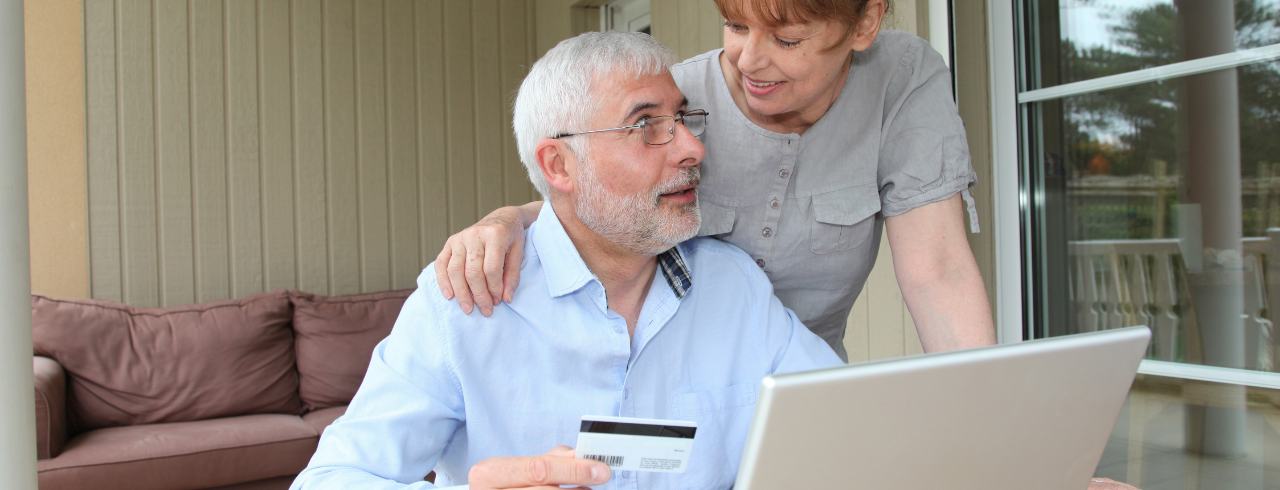 Älterer Herr am PC mit einer Karte in der Hand, eine Frau steht hinter ihm und legt ihm die Hand auf die Schulter