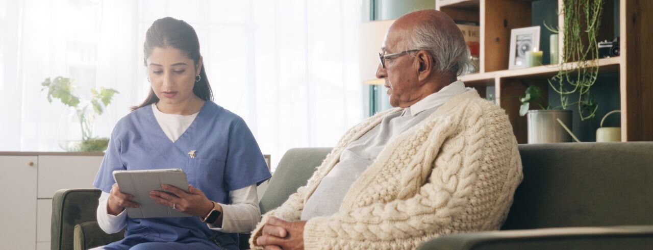 A senior and a caregiver in a living room. The caregiver is holding a tablet.