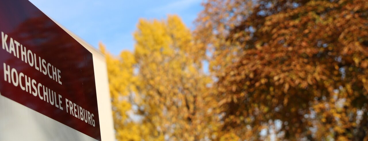 Stele mit der Aufschrift Katholische Hochschule Freiburg vor herbstlichen Laubbäumen und blauem HImmel
