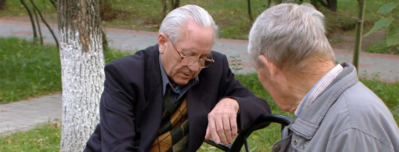 Zwei ältere Herren auf einer Parkbank beschäftigen sich konzentriert mit etwas, das außerhalb des Bildausschnittes liegt.