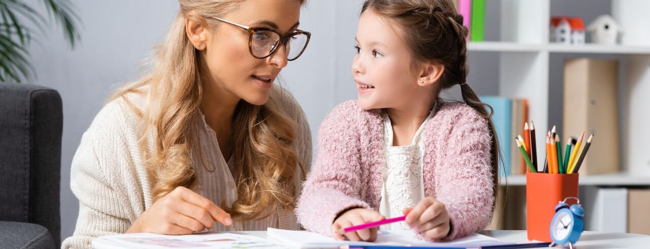 Eine Frau und ein Mädchen sitzen an einem Tisch mit Malsachen vor sich, sie sind miteinander im Gespräch